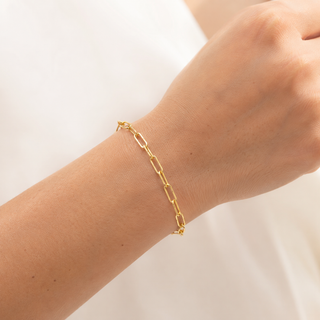 A delicate gold paperclip chain bracelet worn on a person's wrist, shown against a soft white background.